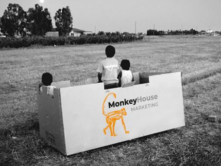 Three children sat in large box in the middle of a feild, box has Monkey House marketing printed on the side. 