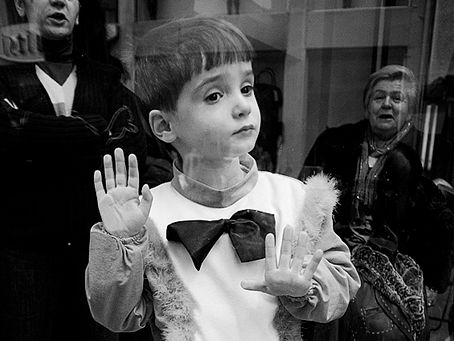 Child in fancy dress with bow tie standing against a window with hand pressed on the glass so it looks as if he in miming